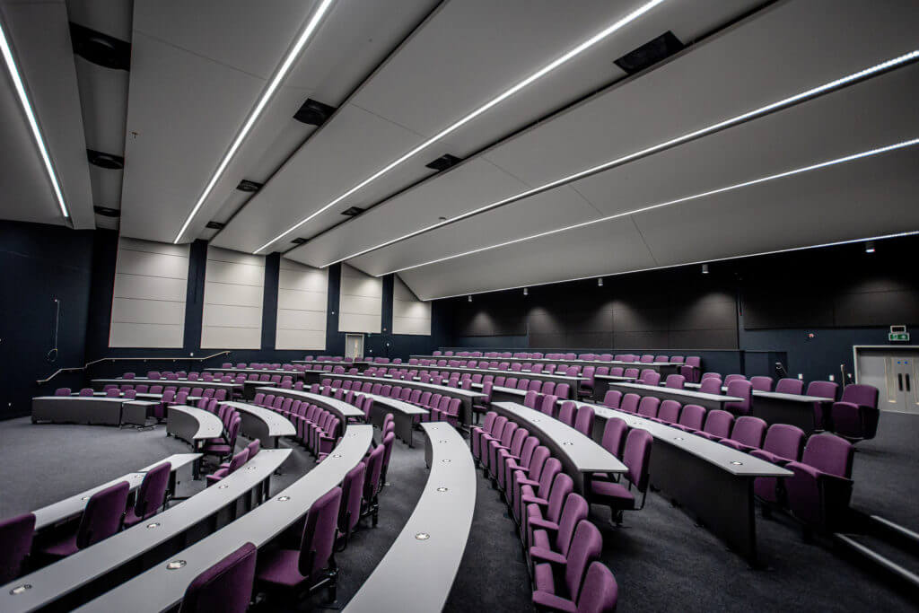 Picture of a lecture hall featuring multiple rows of purple-cushioned chairs.