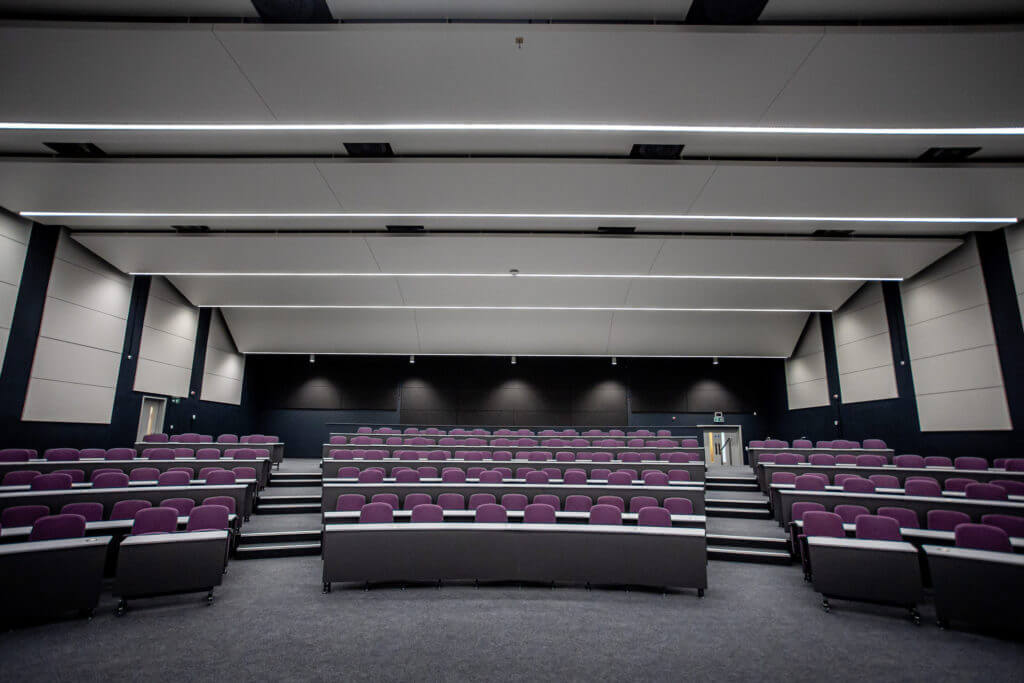 Picture of a lecture hall featuring multiple rows of purple-cushioned chairs.